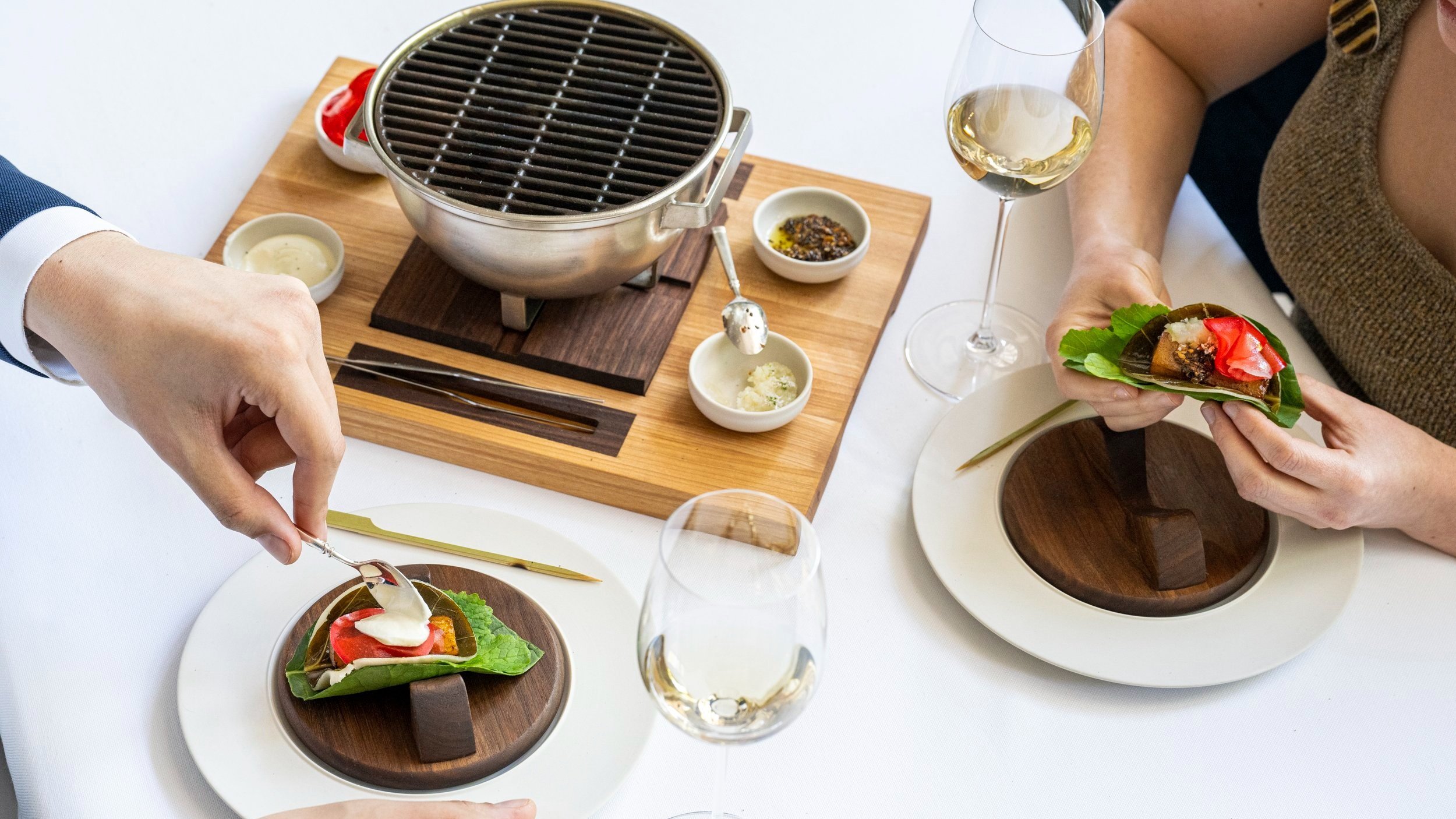 Two people at a dining table with glasses of wine, serving themselves food off of a tabletop grill.